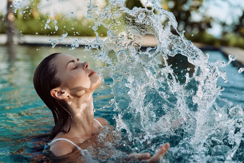Young woman with wet hair enjoys splashing water in a swimming pool on a warm sunny day, expressing relaxation and joy in a casual swimsuit.