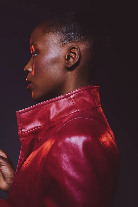 Profile of a young black woman wearing a red leather jacket against a dark background. The image highlights her smooth dark skin and afro hair in a stylish, moody portrait concept.