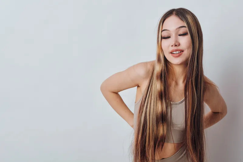 Young woman with long hair wearing casual tank top posed with closed eyes and hands on lower back isolated on light background for beauty care and health support