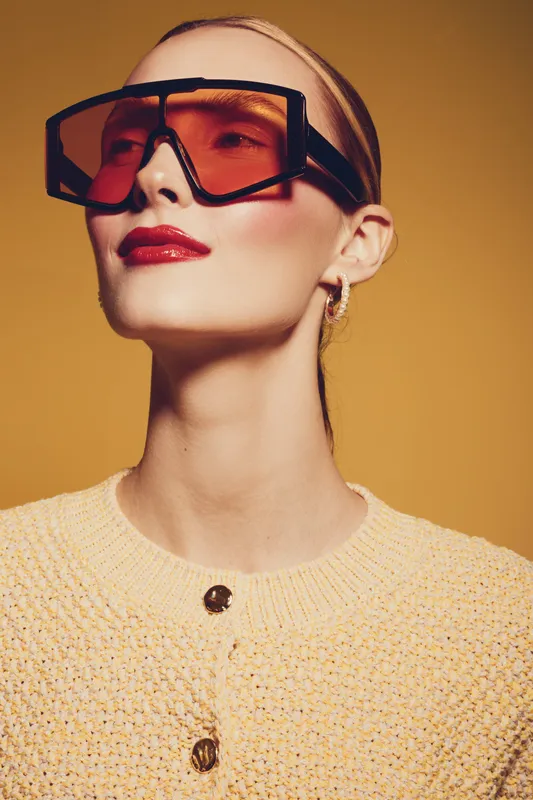 Stylish woman with oversized orange sunglasses and red lipstick poses confidently against warm orange background. Fashion concept with modern accessories and bold makeup.
