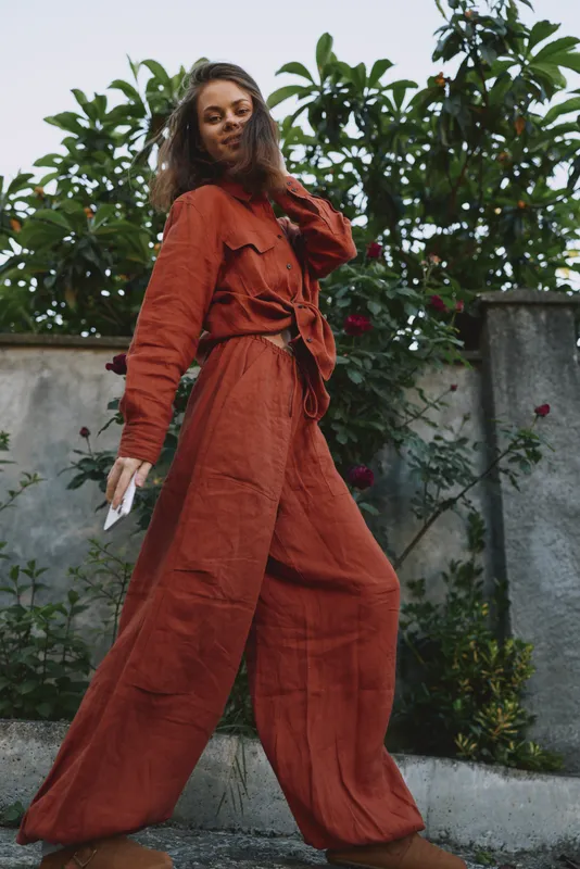 A stylish woman in an orange jumpsuit poses confidently outdoors, belted and wide leg, amid a lush garden backdrop. Fashion lifestyle concept highlighting confidence, color, and outdoor chic
