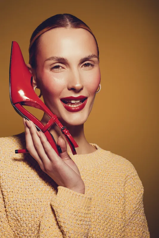 Smiling woman with red lipstick holds red high heel shoe near face, wearing yellow sweater. Studio portrait on brown background. Concept of fashion, beauty, and style accessories.