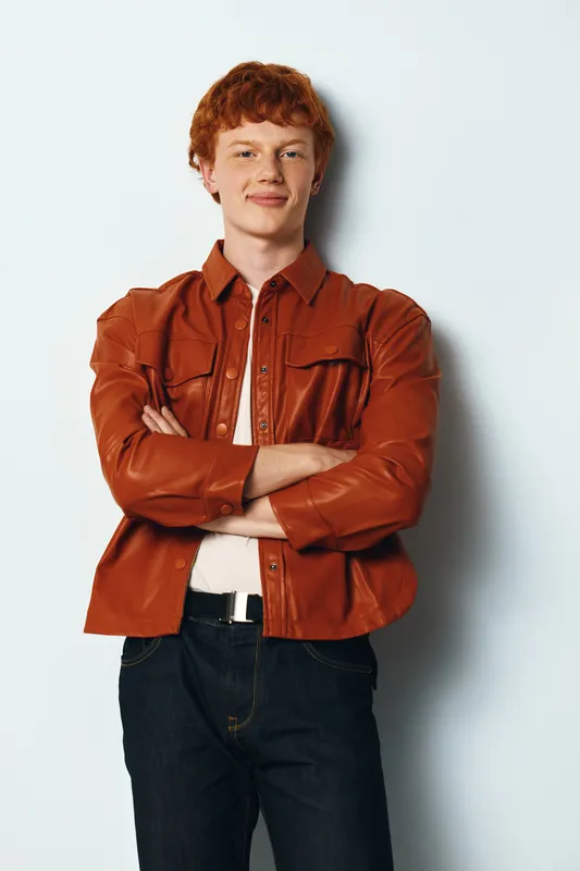 Young man with red hair wearing a brown leather jacket and white shirt standing with arms crossed against white background looking confident.