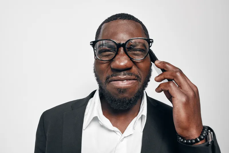 African American man with dark skin wearing glasses and black suit jacket talking on smartphone with expressive face against white background in studio portrait.