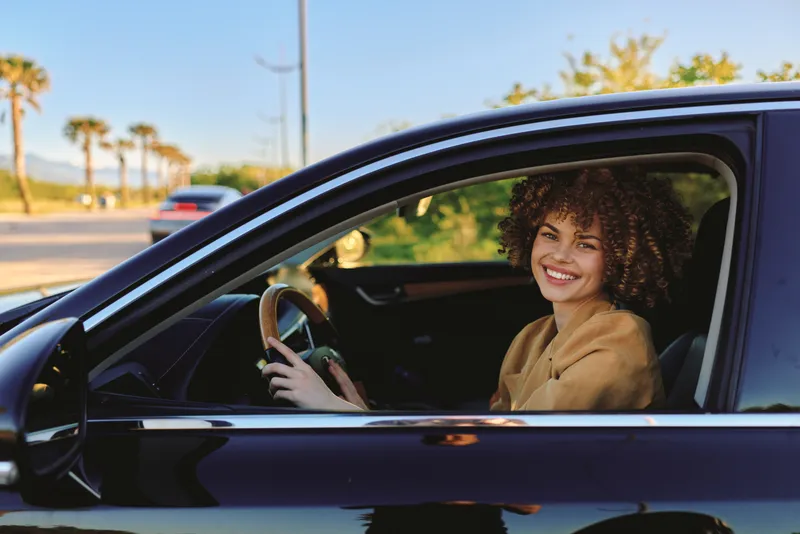 Young woman with curly hair sits in the driver's seat, smiling as she navigates a sunlit street, capturing a moment of everyday independence, casual style, and relaxed confidence behind the wheel.