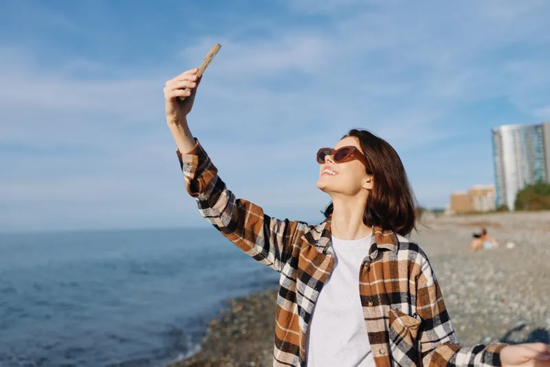Selfie, woman and beach scene with smartphone raised for photo, casual plaid shirt, sunglasses, sunny day, blue sea and horizon, relaxed mood, outdoor portrait for travel, lifestyle and leisure