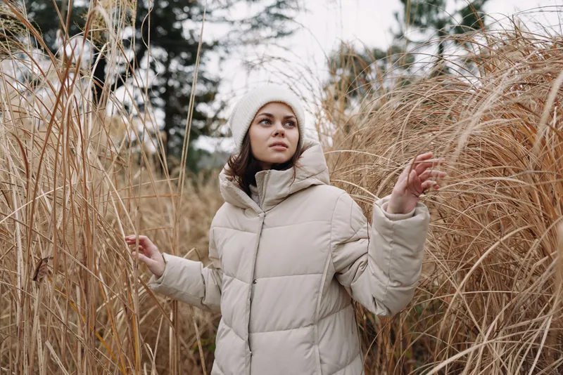 Woman in a warm beige coat stands among tall dried grass in an outdoor winter scene, calm expression, soft light, exploring a quiet natural field.