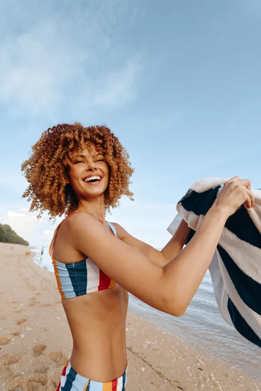 Smiling woman with curly hair in colorful swimsuit holding towel on sunny beach. Clear sky and ocean in background. Concept of summer, vacation, and happiness outdoors.