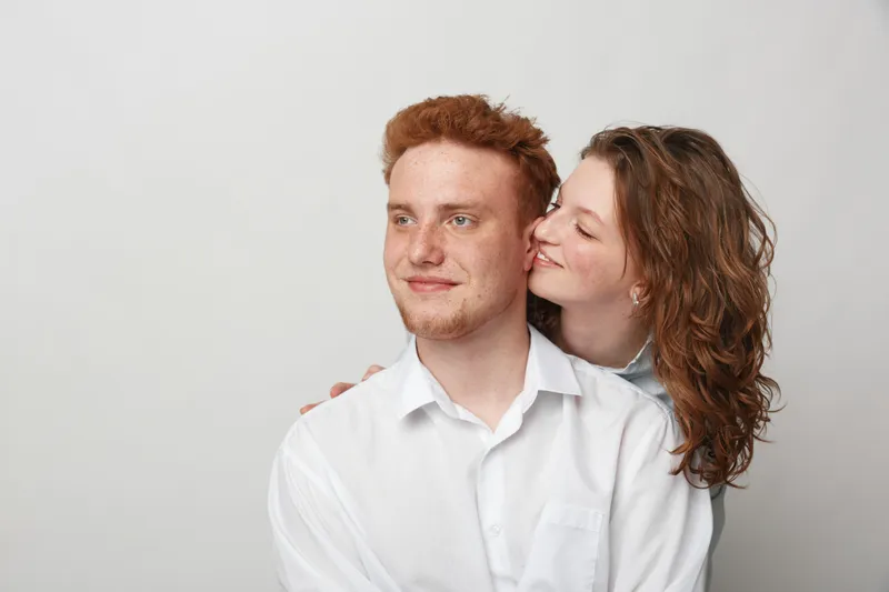 Young couple in white shirts share a tender moment, woman gently kissing man on cheek. Intimate relationship, love, affection, and happiness concept on neutral background.