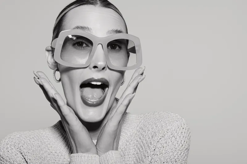Excited white woman with light skin and oversized glasses poses with open mouth and hands near face in studio.