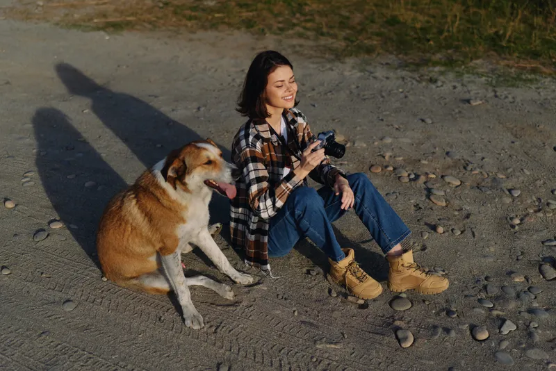 Woman with camera sits on gravel ground beside a dog in an outdoor setting, capturing moments and enjoying a casual hobby under warm light, with relaxed mood and simple nature surroundings.