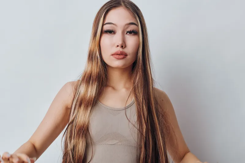 Young woman with long straight hair and natural makeup, wearing a beige tank top, posing in front of a gray background with neutral expression. Beauty, portrait and fashion concept.