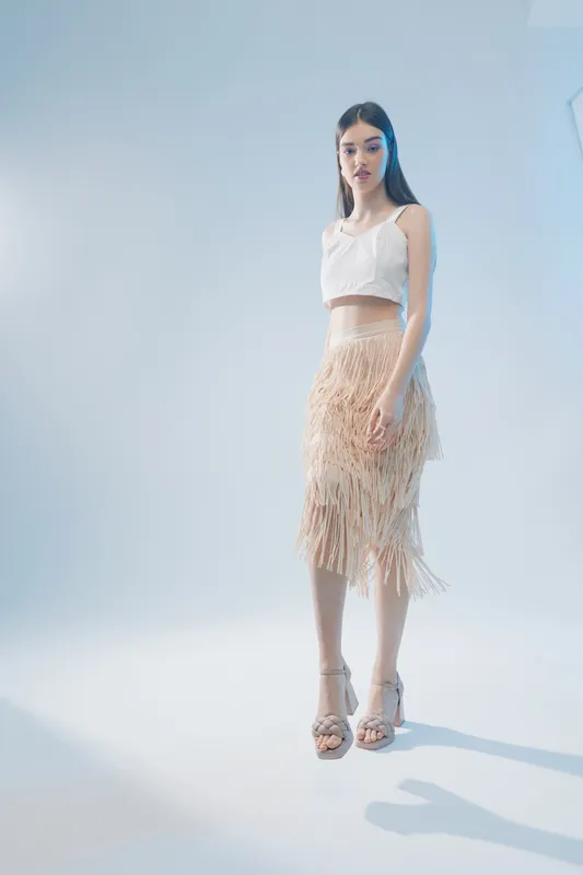 Young woman in a white crop top and beige fringed skirt poses confidently in a bright studio. Concept of woman beauty fashion with natural makeup and sleek hair.