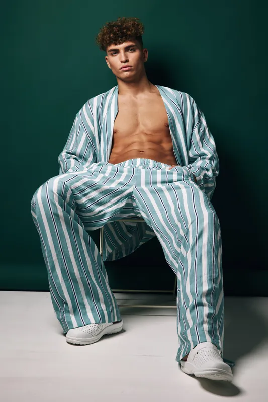 Young man with curly hair posing in striped casual summer outfit with open shirt and wide-leg pants, seated causing relaxed mood fashion style with white sneakers on light background.