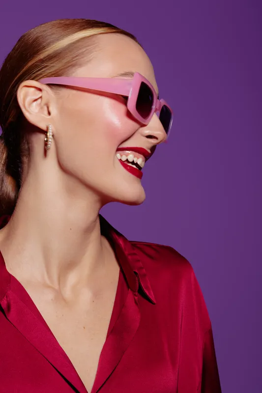Smiling woman with pink sunglasses and red lipstick wears red satin blouse on purple background. Concept of fashion, beauty, style, and confident female portrait in studio lighting.