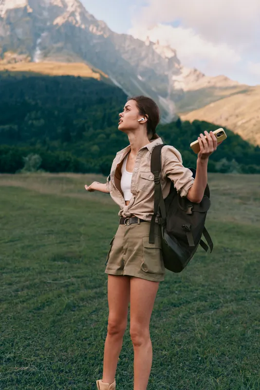Young woman with backpack standing in a lush grassy meadow, towering mountains in the distance, arms raised, holding a book, conveying adventure, outdoor lifestyle, exploration, and fresh air.