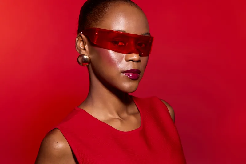 Stylish black woman with dark skin wearing red futuristic sunglasses and red dress poses confidently against a red background. Concept of fashion, beauty, and modern style.