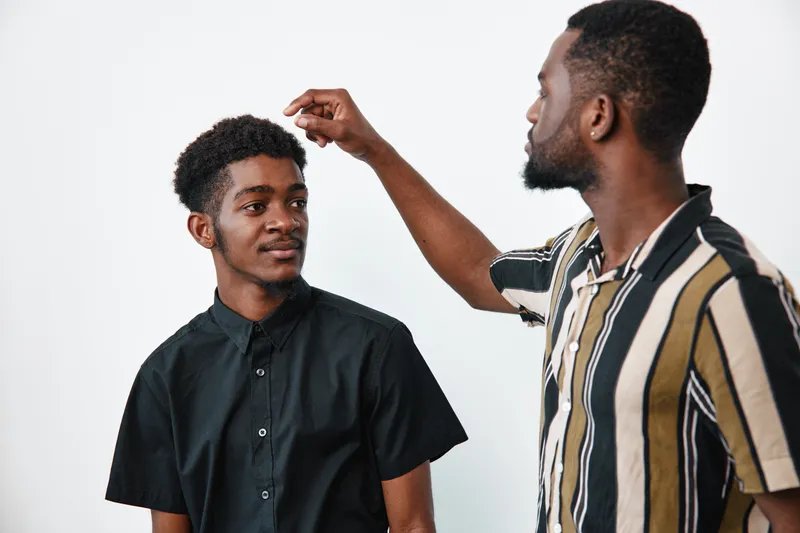 Two African American men with dark skin in casual clothing interacting in studio. One man wearing striped shirt gestures while the other in black shirt listens attentively with slight smile.