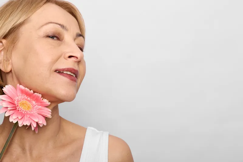 Mature woman, closeup portrait, pink flower near cheek, soft smile, white sleeveless top, light gray background, calm mood, natural makeup, gentle daylight, beauty care routine