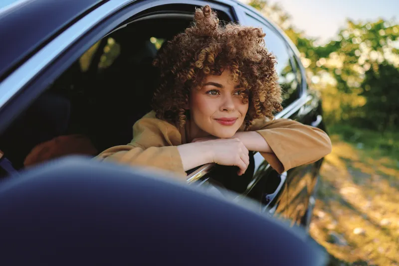 Woman leans from a car window on a sunny outdoor drive, with curly hair and a relaxed smile, capturing a candid lifestyle moment in retro film color
