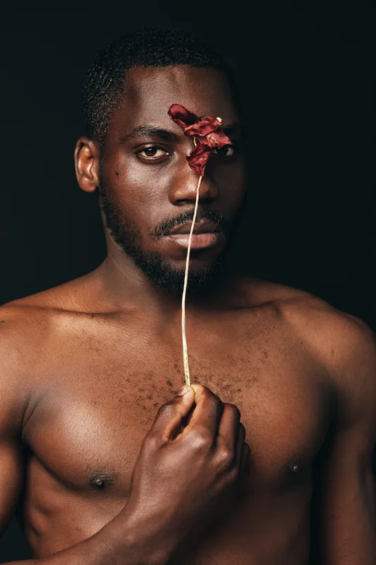 Creative conceptual portrait of African man with bare chest holding dried flower in front of face, dark background, artistic visual style, closeup, emotion and expression.