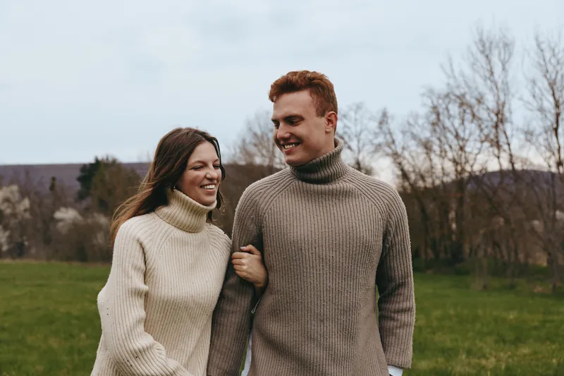 couple, outdoors, sweaters, park, lifestyle, happiness. A smiling couple walks arm in arm through a grassy park, wearing cozy knit sweaters on a cool day, conveying warmth, connection, and relaxed
