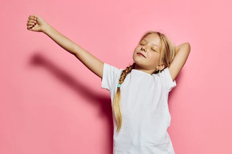 Young girl with long blonde hair stretching arms and closing eyes on pink background. She wears white t-shirt and shows relaxed and peaceful mood. Concept for leisure and childhood.