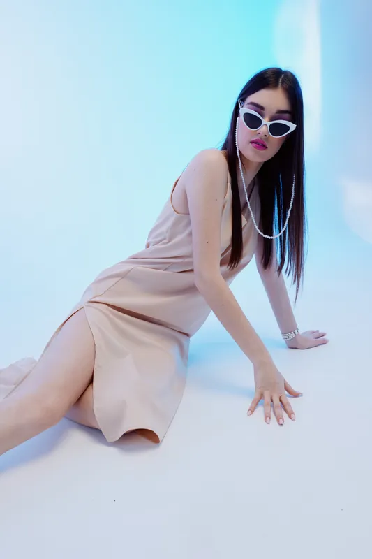 Young woman in a beige dress and white sunglasses poses on the floor in a studio with blue lighting. Concept of beauty, fashion, style, and modern elegance.