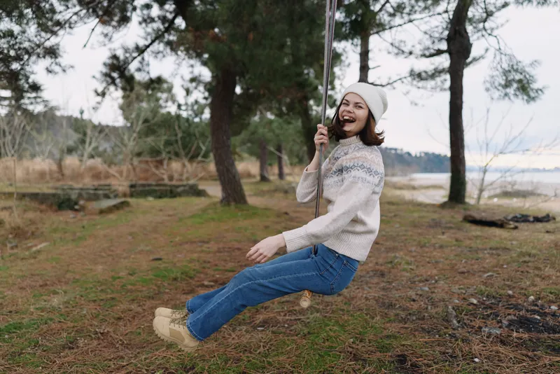 Young woman on a swing in a park by a lake, wearing a knitted sweater and beanie, enjoying a joyful, carefree moment in nature and fresh air.
