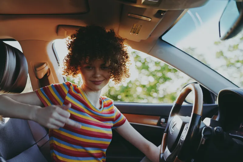 Smiling woman with curly hair wearing a striped shirt sits behind the wheel, capturing a casual lifestyle moment in a warm retro color palette and bright interior.