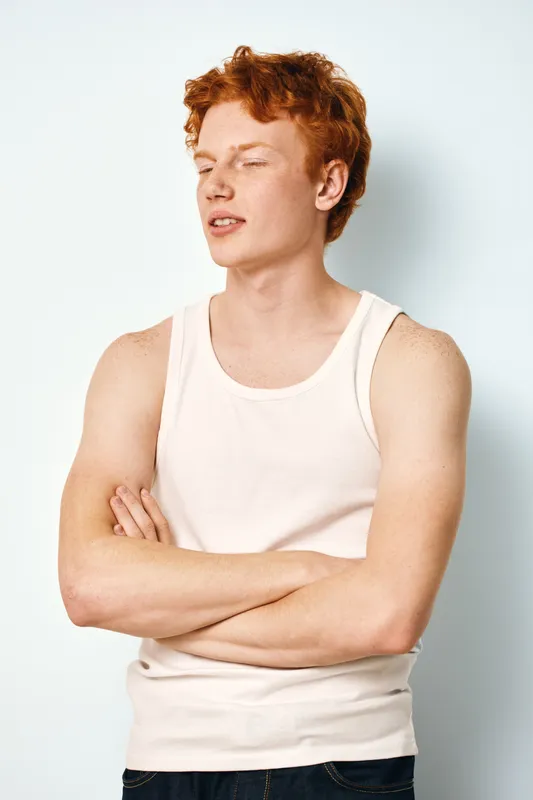 Young man with red hair and freckles wearing white tank top standing with crossed arms against white background. Confident expression and casual style in studio portrait.
