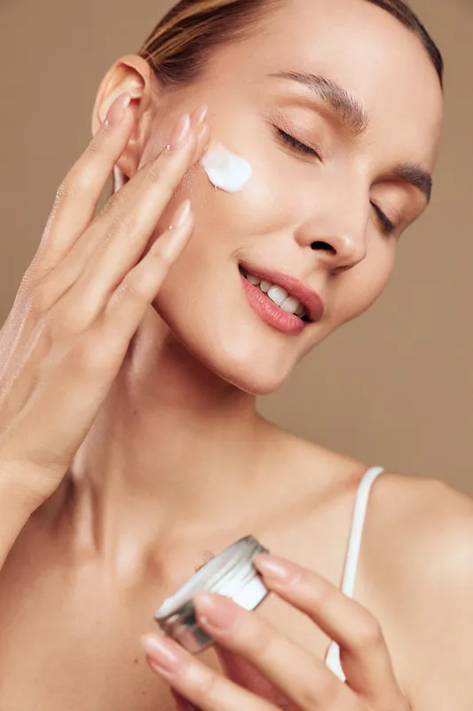 Young woman applying moisturizing cream on face with closed eyes, smooth skin, natural makeup, soft lighting, beige background, skincare routine concept