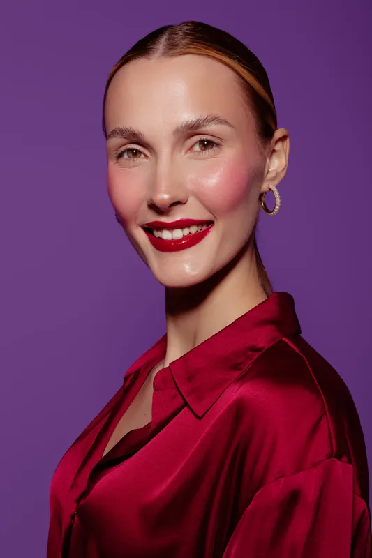 Smiling woman with slicked hair wears red satin blouse and hoop earrings against purple background. Portrait with bright makeup and confident expression. Concept of fashion and beauty.
