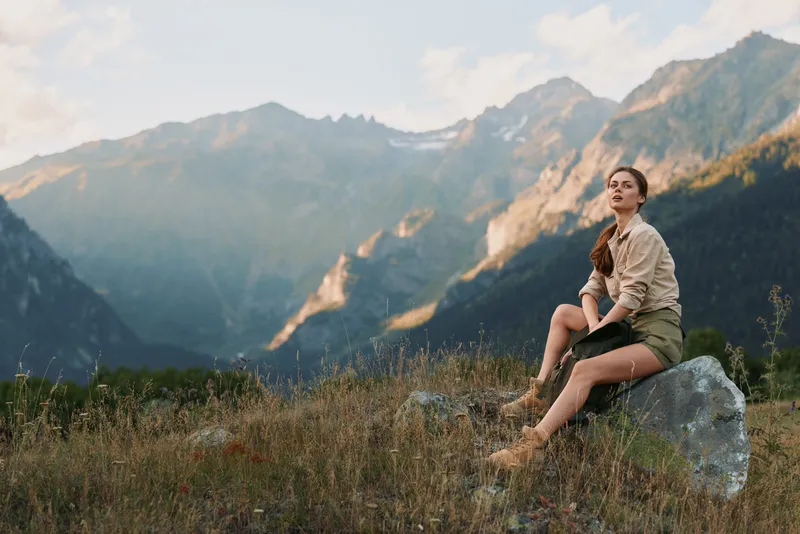Young woman sits on a rock in a mountain meadow relaxed and curious as rugged peaks rise behind her under warm golden light, wearing a light beige shirt and shorts.