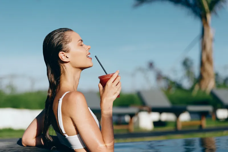 Relaxed woman in a swimsuit sips a red smoothie at poolside, enjoying sunshine, palm trees, and calm water for a carefree summer mood