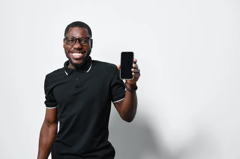African American man with dark skin wearing glasses and black polo shirt smiling and holding smartphone with blank screen on white background.