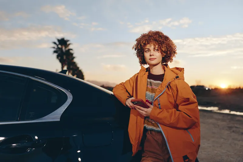 Woman lifestyle, car sunset, travel outdoors, curly jacket fashion. Smiling woman stands by black car on a roadside at dusk, holding jacket, warm golden light, evening sky.