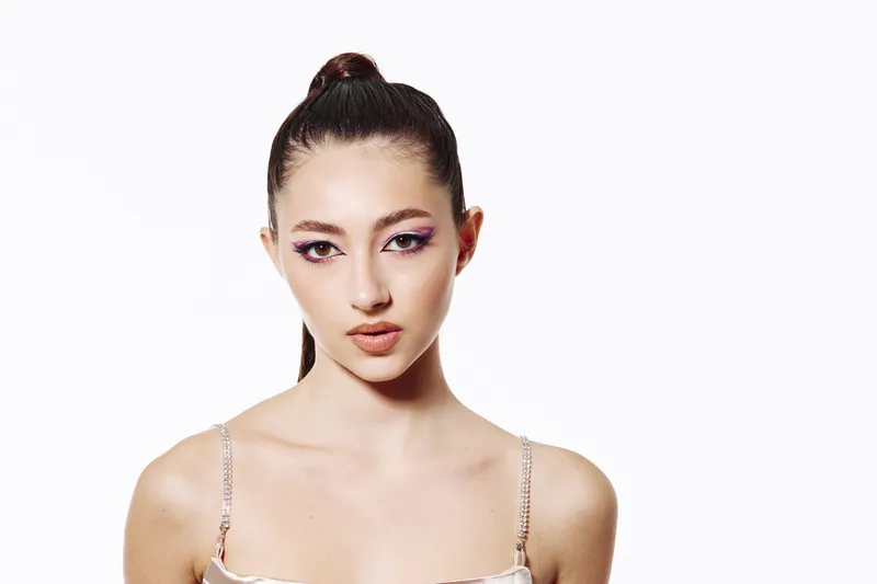 Young woman with sleek topknot hairstyle lines neon purple eyeshadow and nude lipstick against white background. Creative fashion beauty portrait with modern subtle glow concept.
