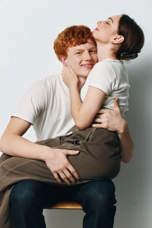 Couple, young man and woman embracing, smiling and sitting close together on chair for love, affection and relationship in casual clothing on plain background.