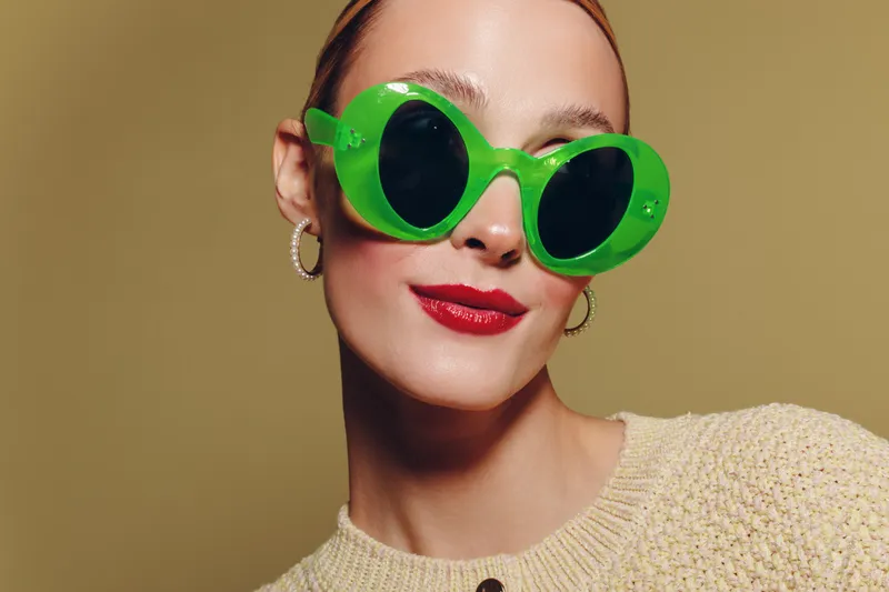 Stylish woman wearing large green sunglasses and red lipstick poses confidently against beige background. Fashion concept with bold accessories and modern makeup look.