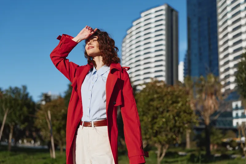 Young woman with curly hair enjoys sunny weather while wearing a red coat and light trousers, standing outdoors near modern buildings with a relaxed and content expression.