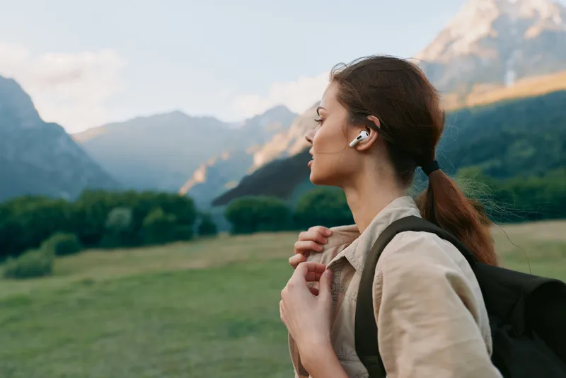 Woman with backpack in a scenic meadow, wearing wireless earbuds, gazing at distant mountains, capturing a vivid outdoor lifestyle moment of exploration and wellness, outdoors