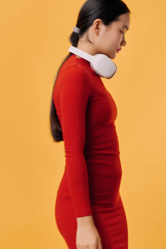 Young woman with long hair wearing red dress and white headphones standing sideways on orange background. Profile portrait of serious girl in modern casual style with minimalistic setting for fashion.