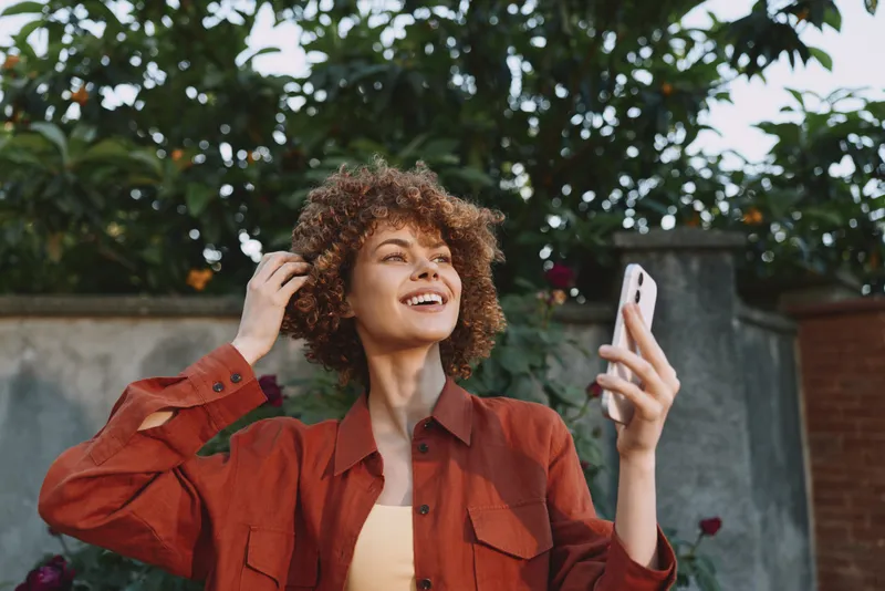 A cheerful young woman with curly hair smiles as she selfies outdoors, wearing a rust red jacket while standing among leafy greenery and a sunlit garden
