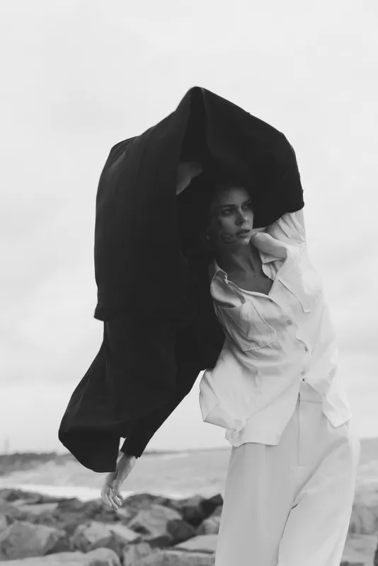 Fashion on the shore shows a model in flowing white garments as a heavy cloak billows overhead, capturing wind, movement, and dramatic contrast between soft fabric and stark sky.