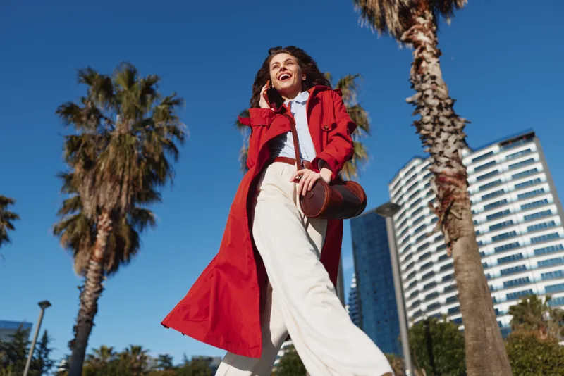 Young woman walking outdoors wearing red coat and white pants, smiling and talking on phone, sunny day, palm trees and modern buildings in background