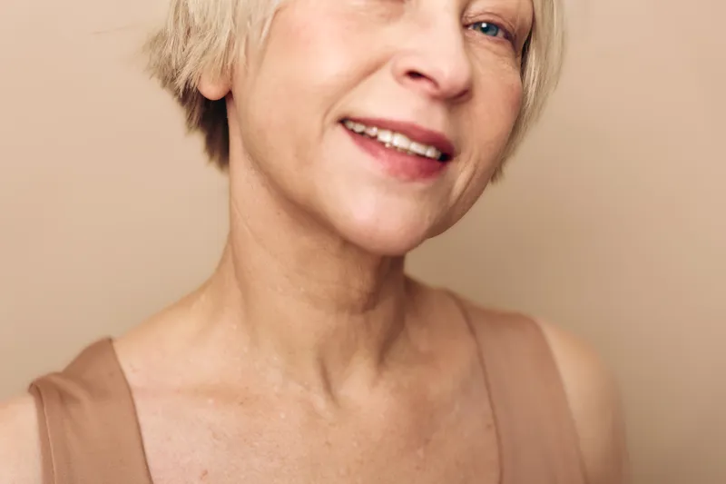 Close up of smiling mature woman with short blonde hair and natural skin in beige top, isolated on light neutral background, concept of aging gracefully and natural beauty.