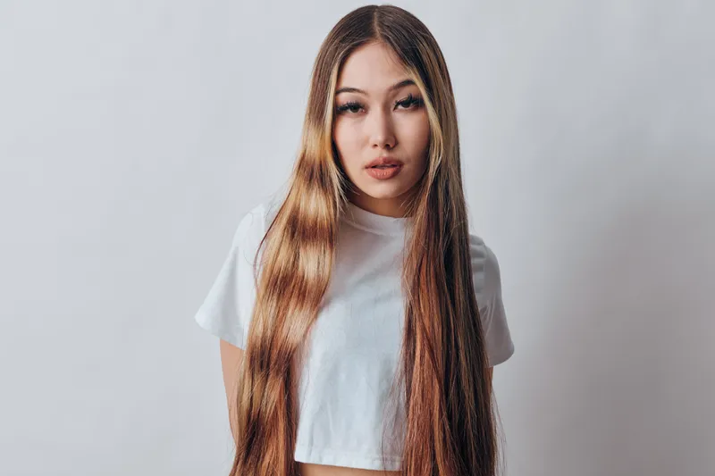 Young woman with long straight hair and neutral expression wearing white t shirt standing against light background in studio for fashion beauty portrait and hairstyle concept.
