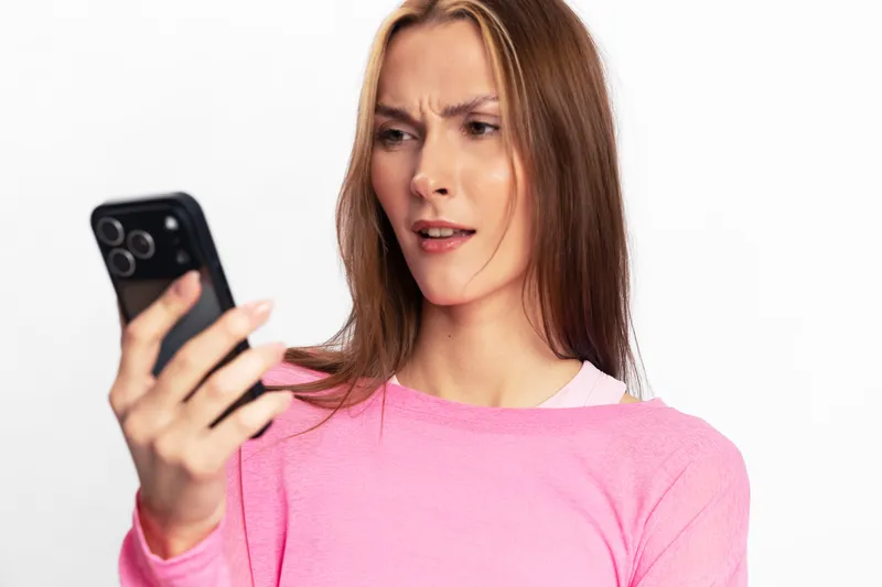 Confused young woman in pink sweater looks at smartphone with puzzled expression isolated on white background. Concept for misunderstanding, surprise, or unexpected information on mobile device.