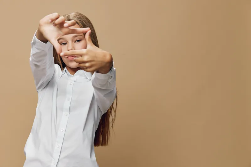 Girl with long hair and white shirt framing face with hands in focus for creativity, vision and photography concept on beige background with casual look and serious expression.